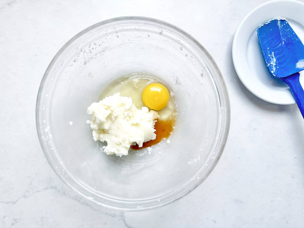A glass bowl contains an egg, sugar, and vanilla extract, ready for mixing. A blue spatula with some residue rests on a small white plate nearby.