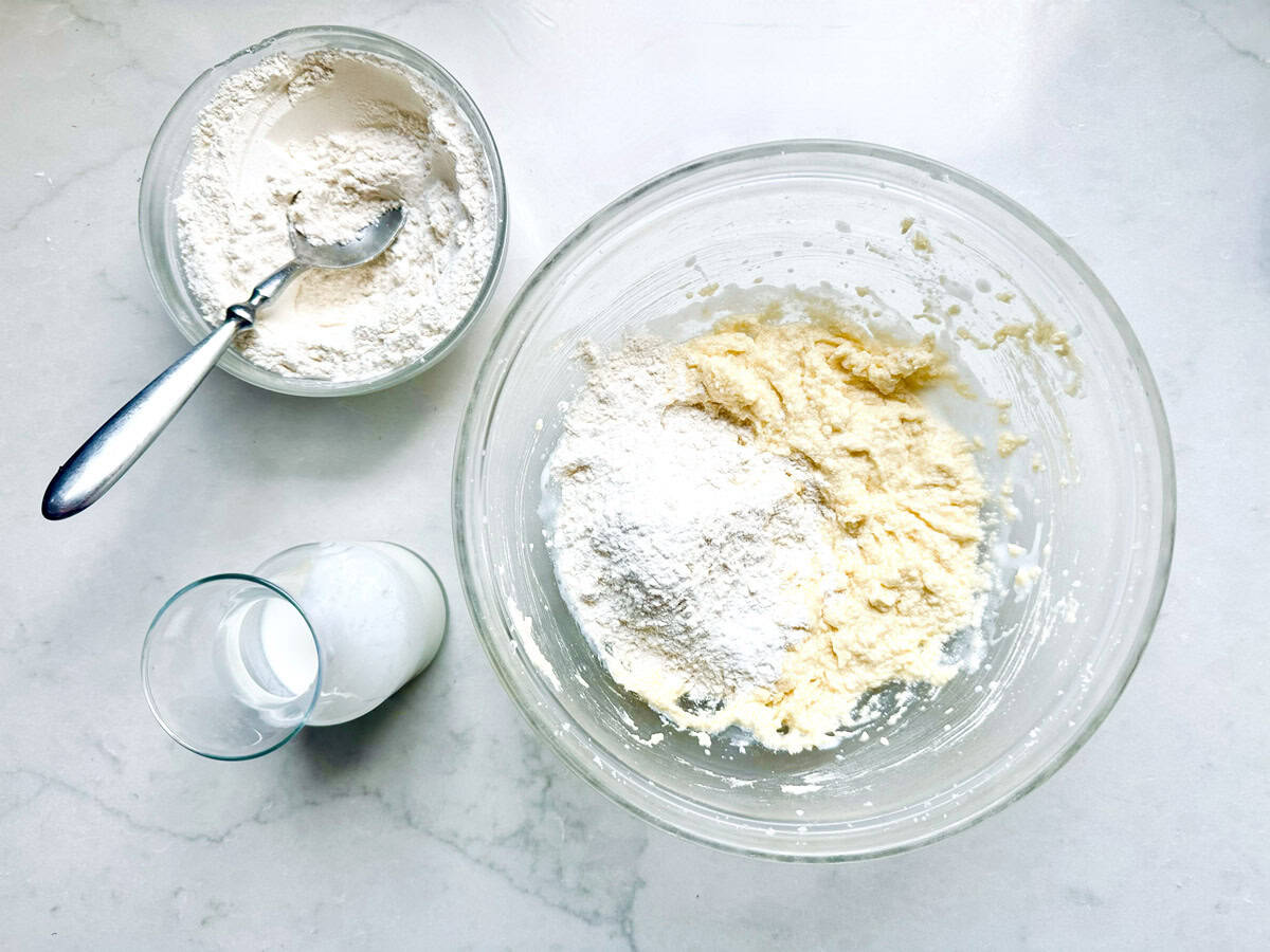 A glass bowl with flour and dough, a bowl of flour with a spoon, and a glass of milk are on a white countertop.