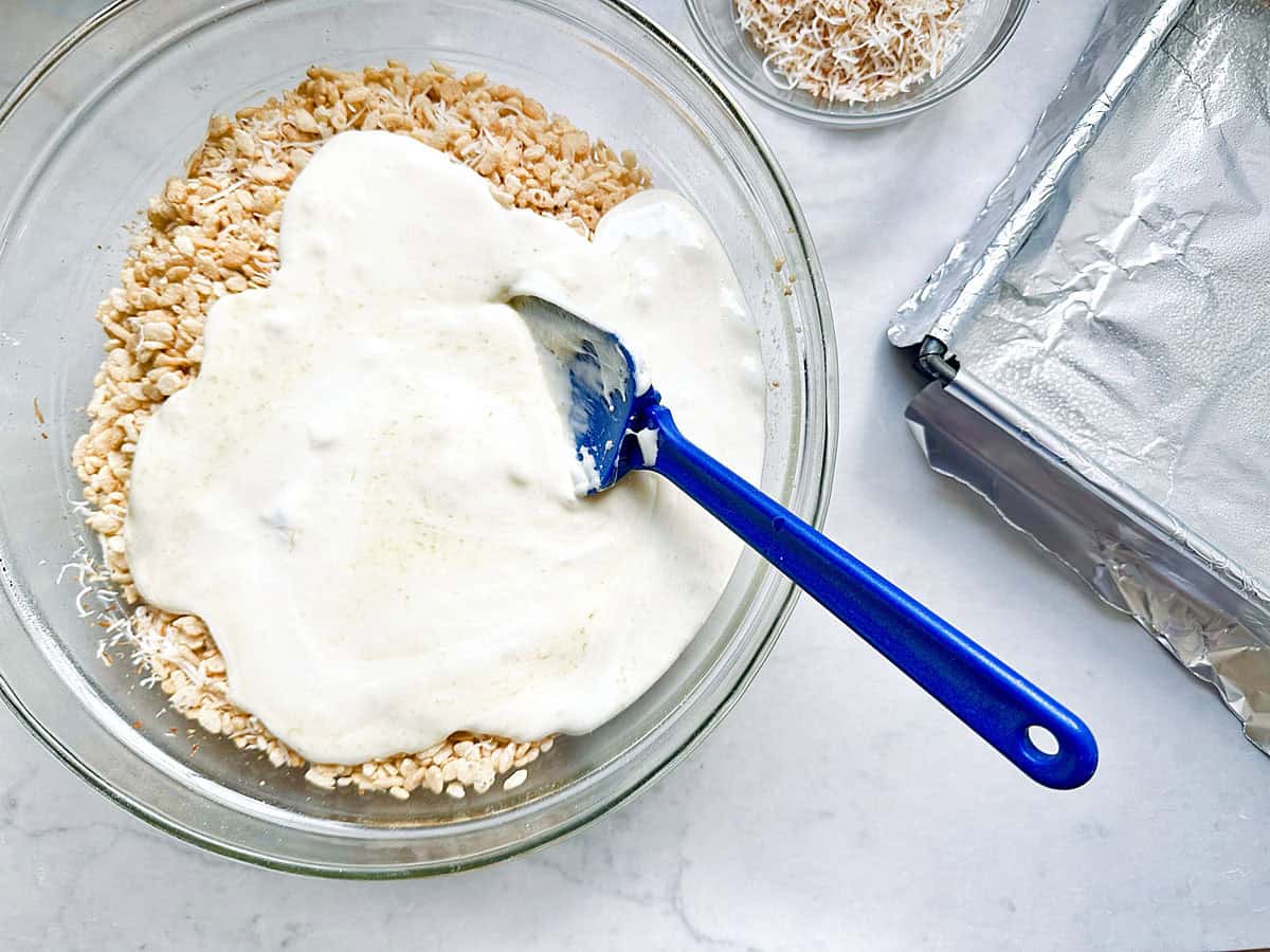 A bowl of rice cereal topped with melted marshmallow mixture is being mixed with a blue spatula. Next to it are a small bowl of toasted coconut and a foil-lined baking pan.