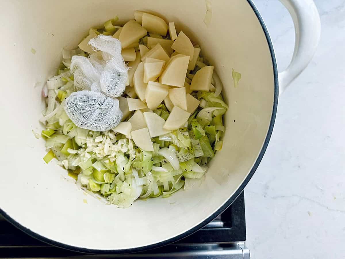 Diced potatoes, chopped leeks, onions, and a bundle of herbs in cheesecloth are in a white pot on a stovetop, ready to be cooked into a comforting Potato Leek Soup.