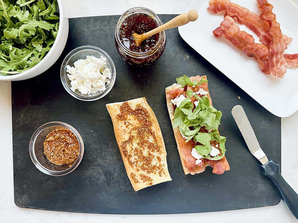 A Bacon Fig and Arugula Sandwich is being assembled with ciabatta bread, crispy bacon, fresh arugula, and creamy goat cheese on a black cutting board. Nearby are a jar of fig jam, a bowl of arugula, a dish with bacon, and a bowl of mustard.