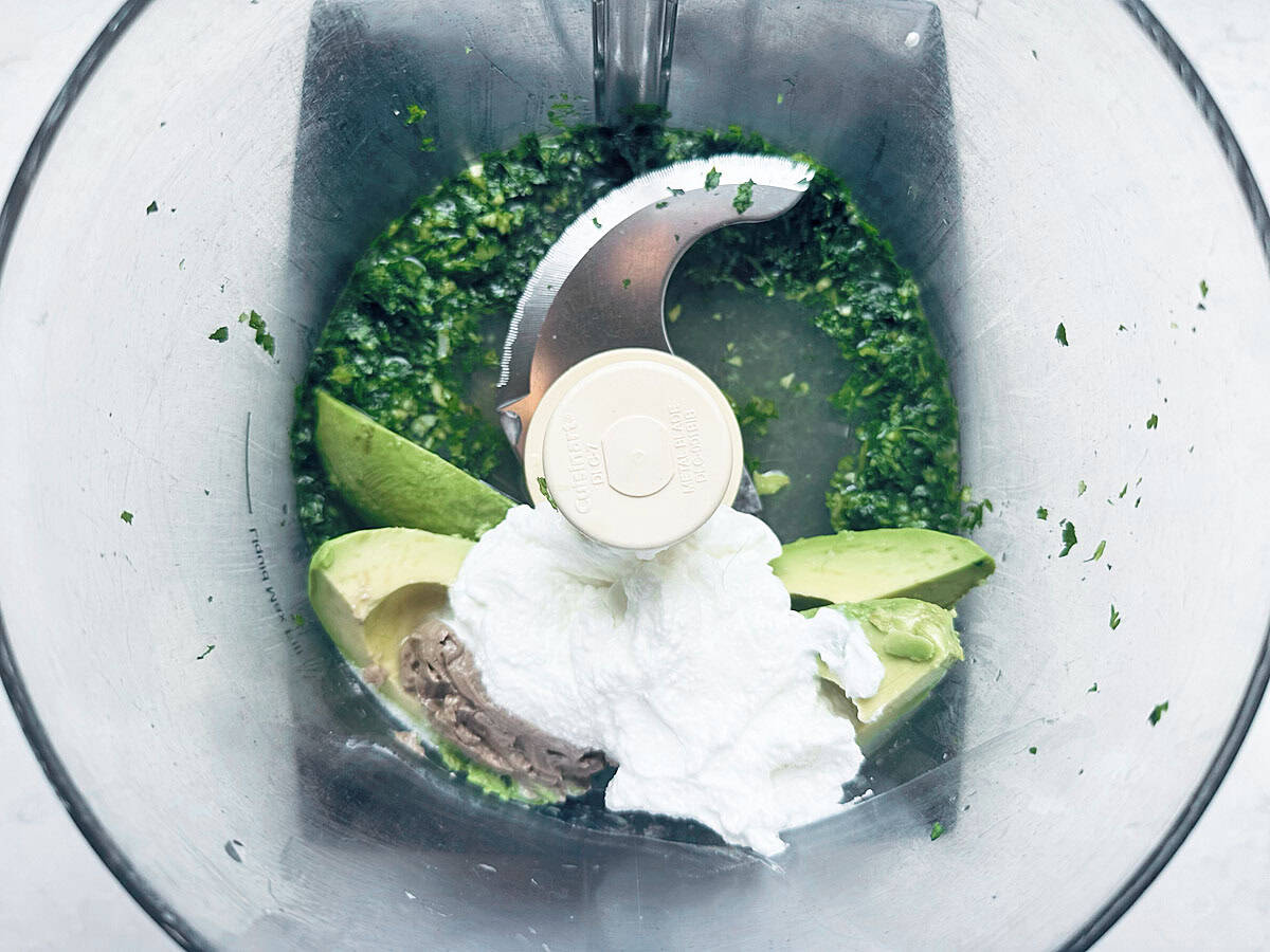 Overhead view of a food processor bowl containing chopped herbs, avocado slices, a dollop of yogurt, and a sharp blade in the center, ready for blending.