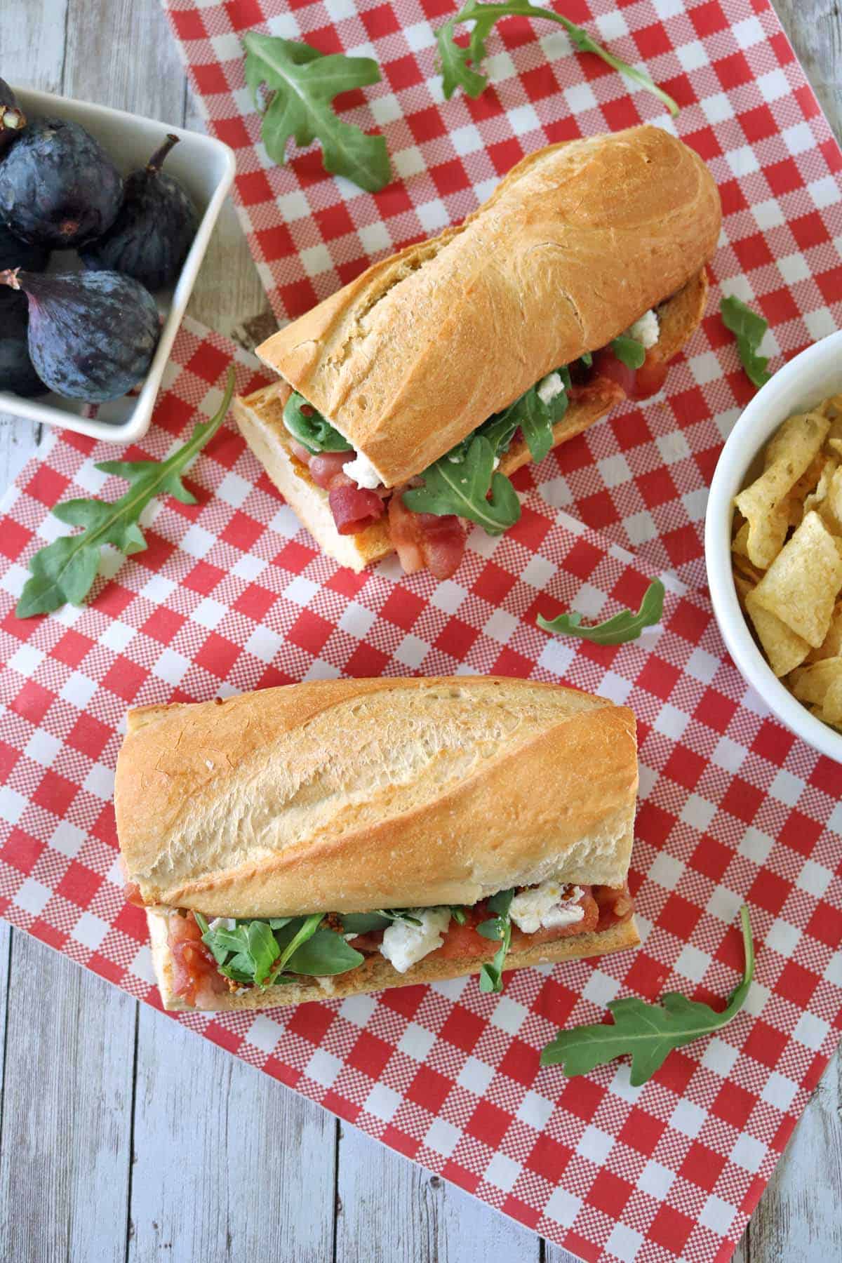 Two baguette sandwiches with arugula and cheese on a red checkered cloth, next to a bowl of tortilla chips and a dish of fresh figs on a wooden table.