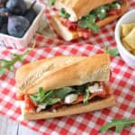 A baguette sandwich with arugula, cheese, and figs sits on a red checkered cloth. Nearby are a bowl of figs and a bowl of chips.
