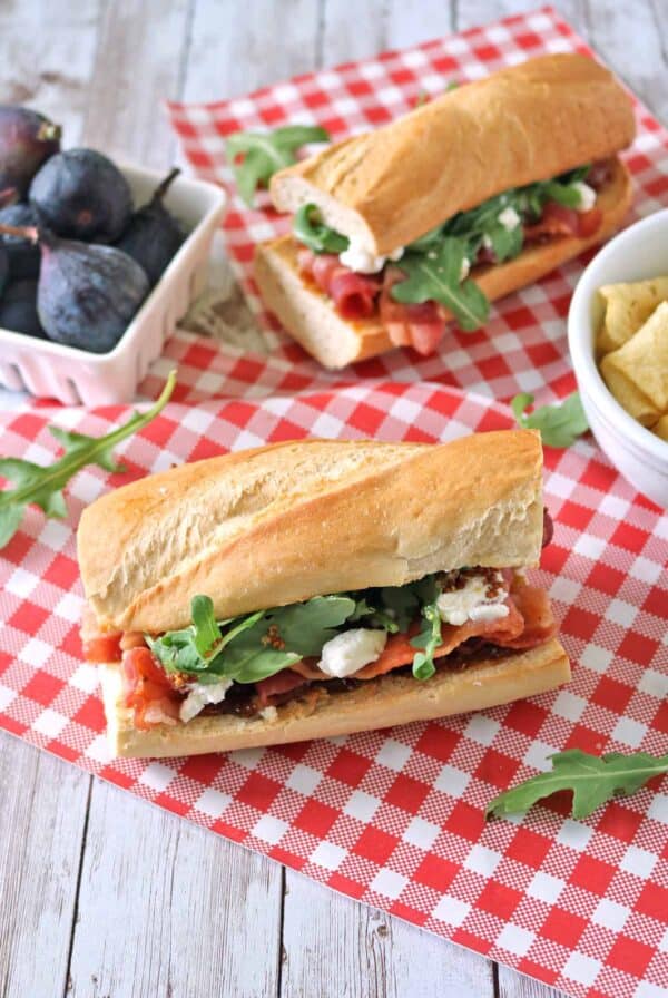 A baguette sandwich with arugula, cheese, and figs sits on a red checkered cloth. Nearby are a bowl of figs and a bowl of chips.
