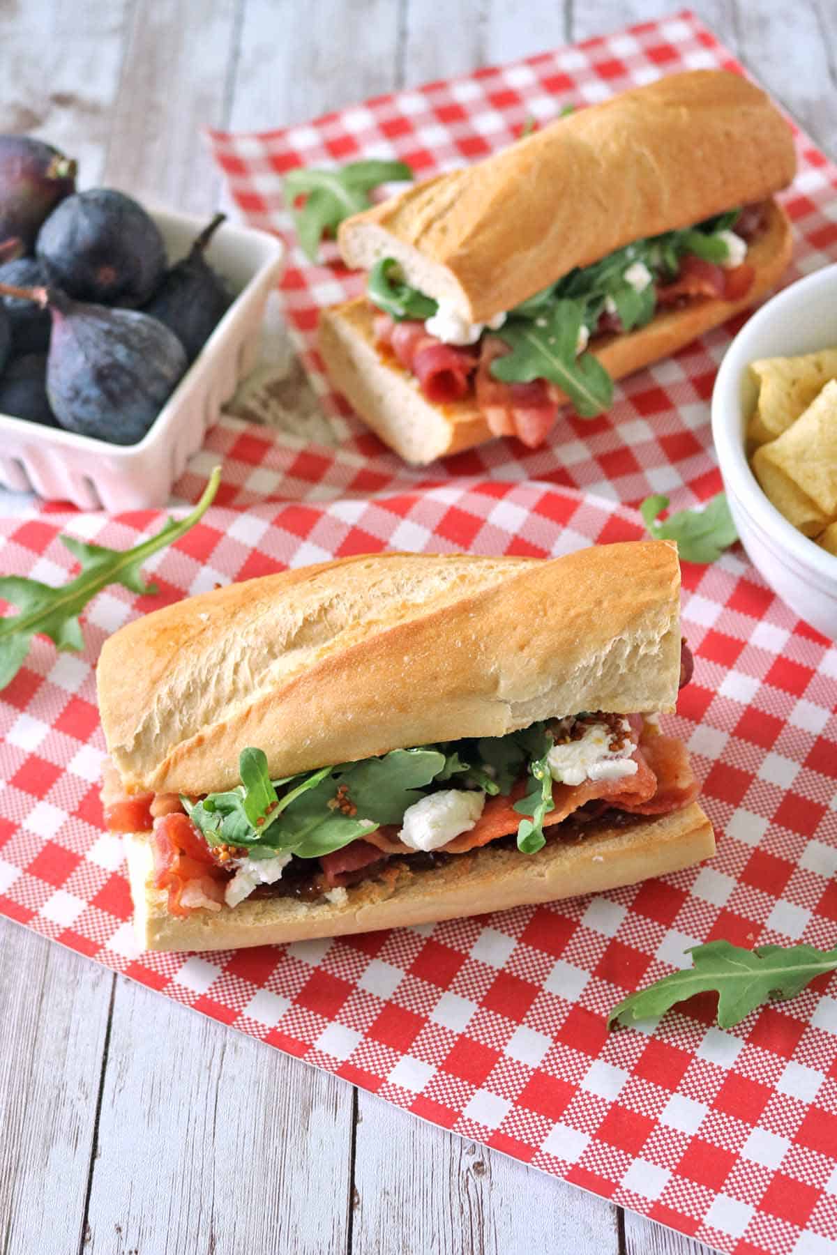 A baguette sandwich with arugula, cheese, and figs sits on a red checkered cloth. Nearby are a bowl of figs and a bowl of chips.