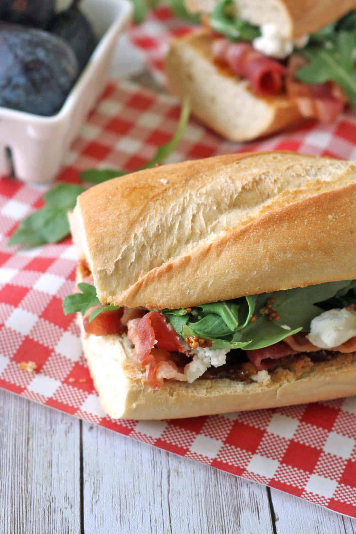 A sandwich with lettuce, prosciutto, and cheese on a baguette sits on a red and white checkered cloth, with a container of figs in the background.