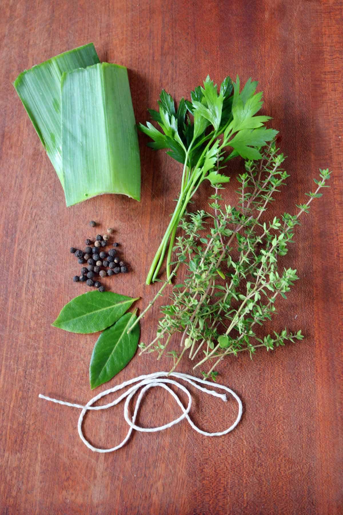 Fresh herbs (parsley, thyme, bay leaves), black peppercorns, leeks, and kitchen twine are arranged on a wooden surface, demonstrating how to make a bouquet garni and sachet for your favorite soups and stews.