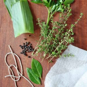 A bunch of herbs, bay leaves, black peppercorns, string, cheesecloth, and leek leaves are arranged on a wooden surface—classic ingredients you’ll need if you’re wondering how to make a bouquet garni and sachet.