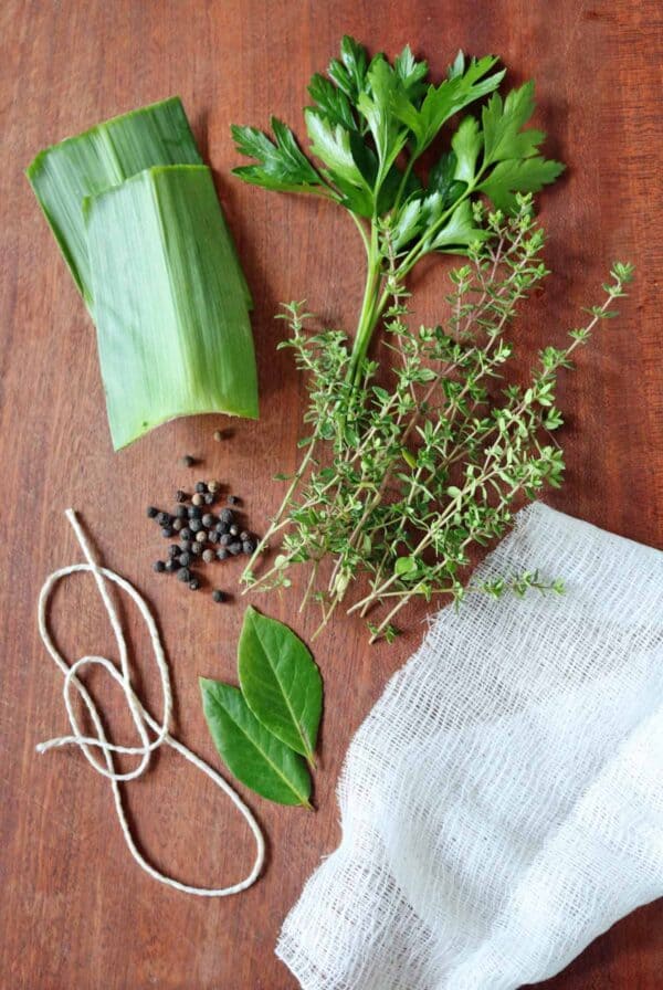 A bunch of herbs, bay leaves, black peppercorns, string, cheesecloth, and leek leaves are arranged on a wooden surface—classic ingredients you’ll need if you’re wondering how to make a bouquet garni and sachet.