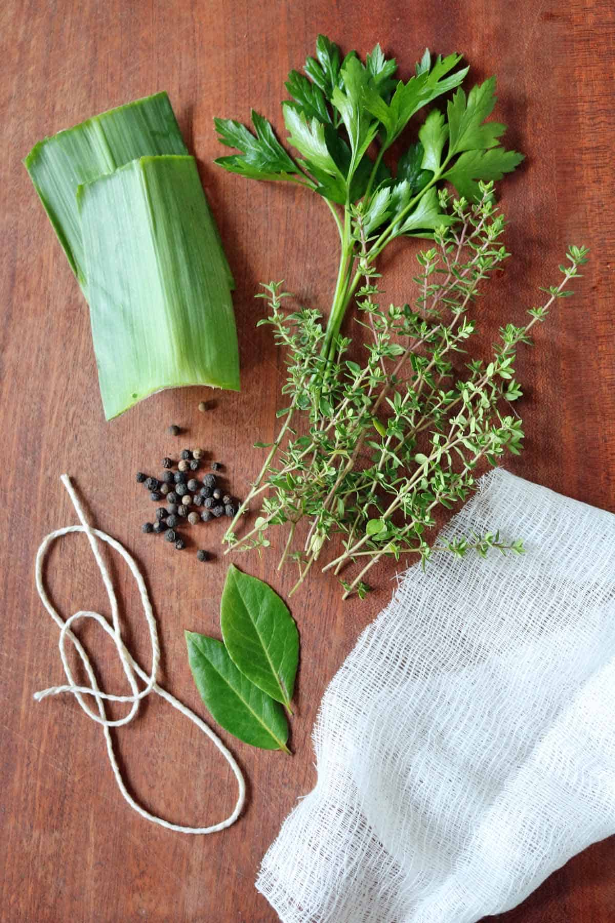 A bunch of herbs, bay leaves, black peppercorns, string, cheesecloth, and leek leaves are arranged on a wooden surface—classic ingredients you’ll need if you’re wondering how to make a bouquet garni and sachet.