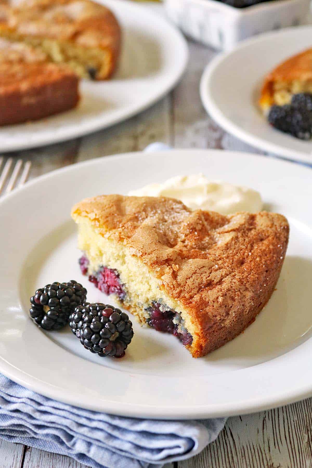 A slice of berry cake with visible blackberries, served on a white plate with fresh blackberries and a dollop of cream on the side.