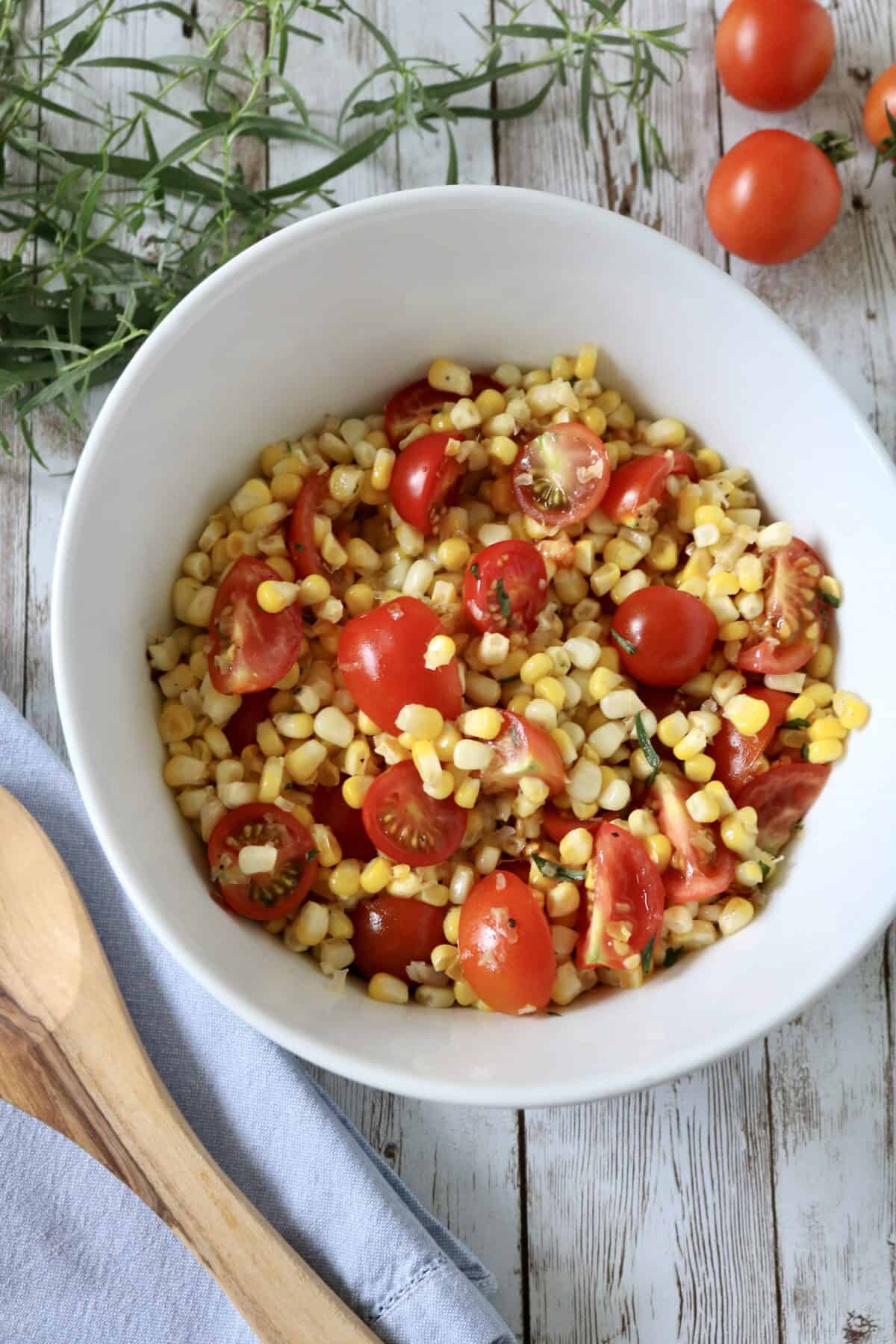 A white bowl filled with corn, cherry tomato halves, and herbs sits on a wooden table next to a wooden spoon, sprigs of fresh herbs, and loose cherry tomatoes.