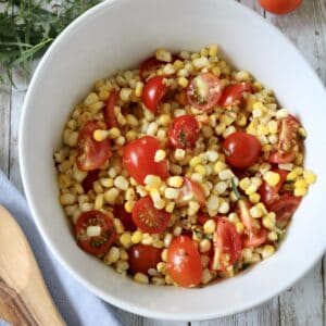A white bowl filled with corn, cherry tomato halves, and herbs sits on a wooden table next to a wooden spoon, sprigs of fresh herbs, and loose cherry tomatoes.