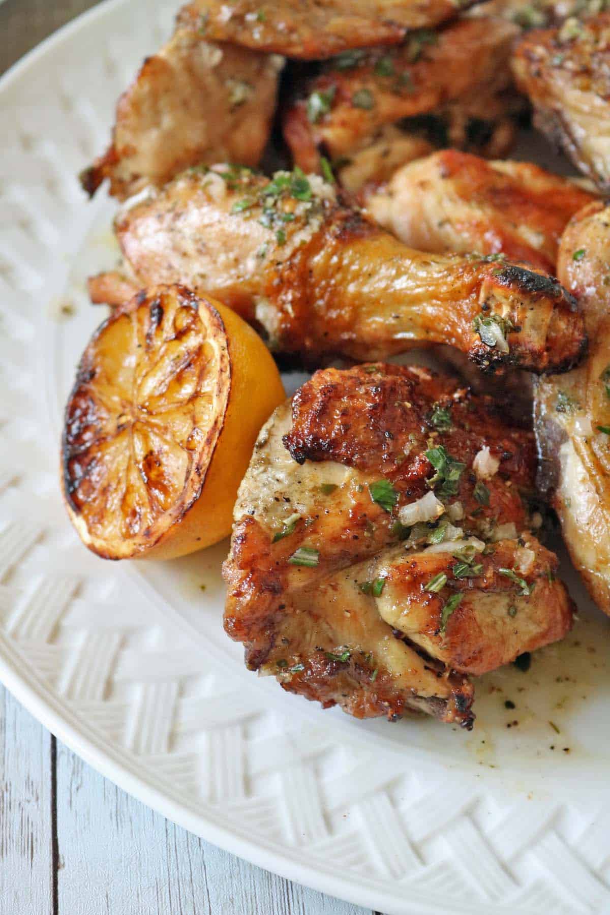 Grilled chicken pieces garnished with herbs served on a white plate, accompanied by a charred lemon half. The chicken has visible grill marks and a golden-brown, crispy exterior.