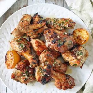 A white plate filled with grilled chicken drumsticks and wings, garnished with herbs and served with charred lemon halves. A cup of green sauce and a patterned napkin are nearby on a rustic wooden table.
