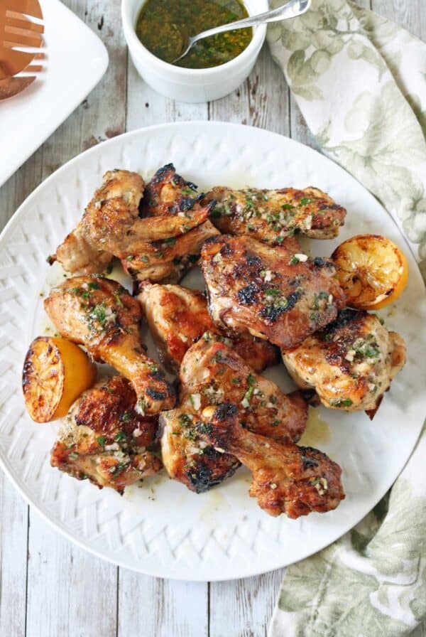 A white plate filled with grilled chicken drumsticks and wings, garnished with herbs and served with charred lemon halves. A cup of green sauce and a patterned napkin are nearby on a rustic wooden table.