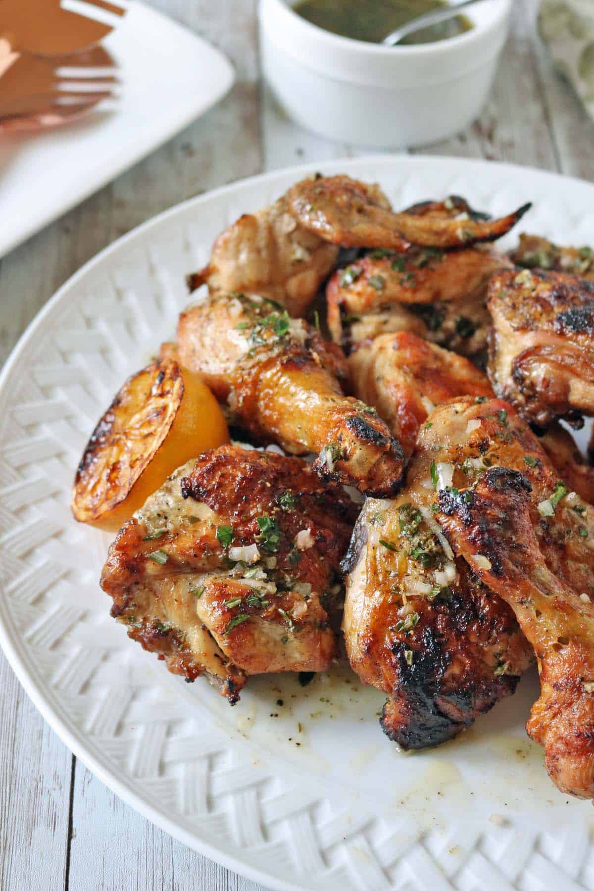 Grilled chicken wings garnished with herbs on a white plate, served with a roasted lemon wedge. A bowl of green dipping sauce is in the background on a rustic wooden table.