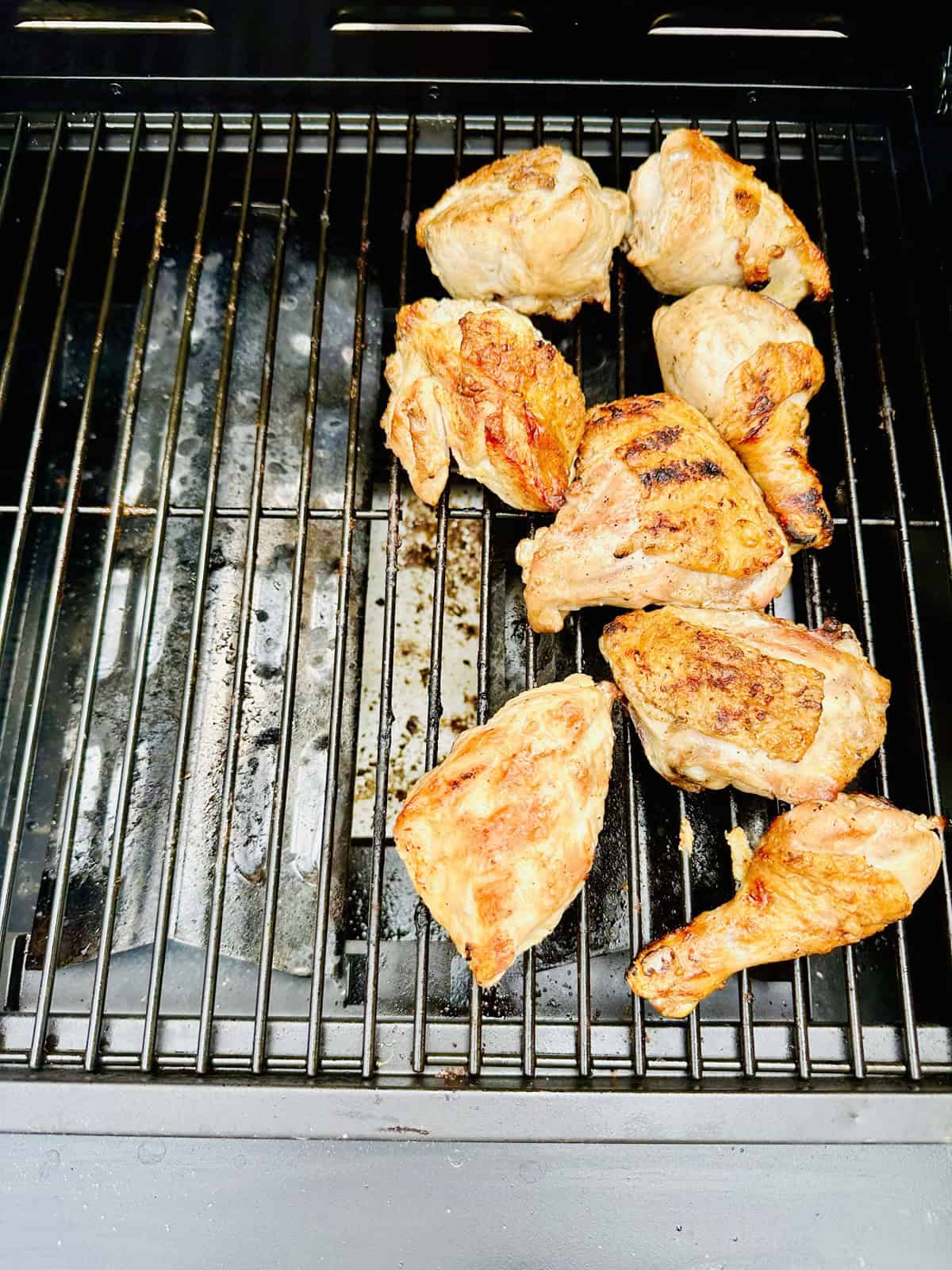Grilled chicken pieces cooking on an outdoor grill, some with visible grill marks, arranged on the right side of the grates.
