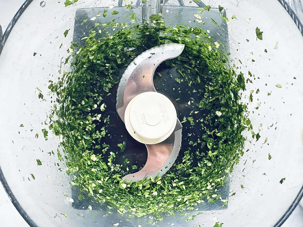 Chopped green herbs in a food processor, viewed from above, with metal blades visible in the center of the clear container.