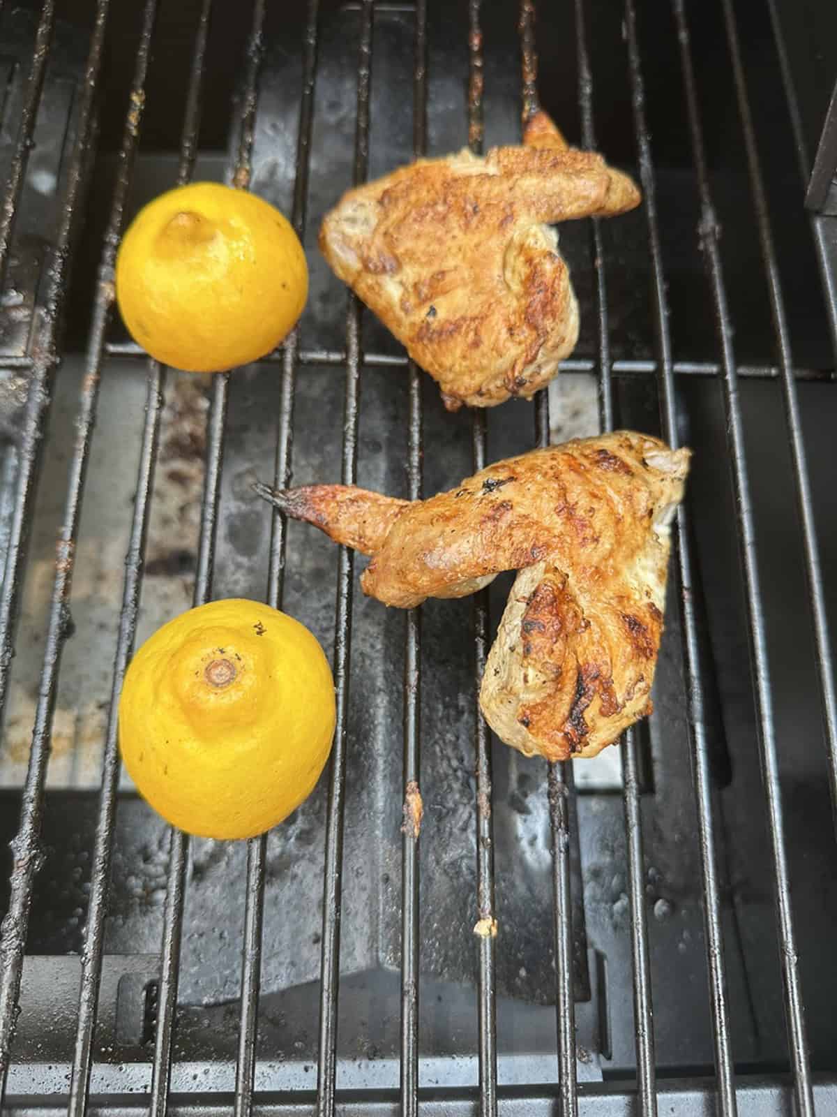 Two grilled chicken wings and two halved lemons are cooking on a barbecue grill, with char marks visible on the chicken and lemons.