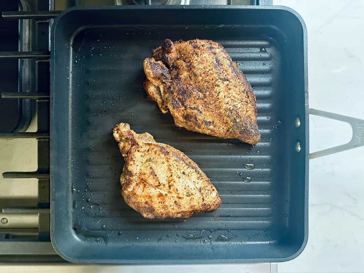 Two seasoned chicken breasts cooking on a black stovetop grill pan, with grill marks visible and some oil droplets on the pan surface—perfect for assembling into flavorful Grilled Street Corn Chicken Bowls.