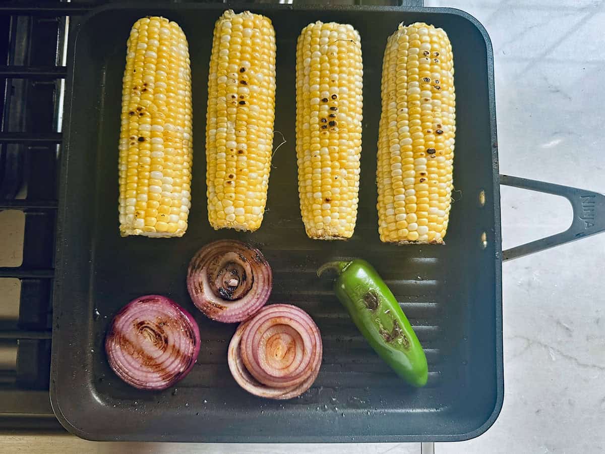 Four ears of corn, two red onion slices, and one green jalapeño are being grilled on a black grill pan, showing light char marks on the vegetables—the perfect start for flavorful Grilled Street Corn Chicken Bowls.