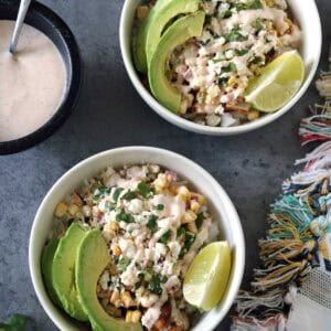 Two bowls of Grilled Street Corn Chicken Bowls topped with avocado slices, lime wedges, cheese, and creamy dressing sit on a gray surface, accompanied by a colorful cloth, fresh cilantro, and extra dressing on the side.
