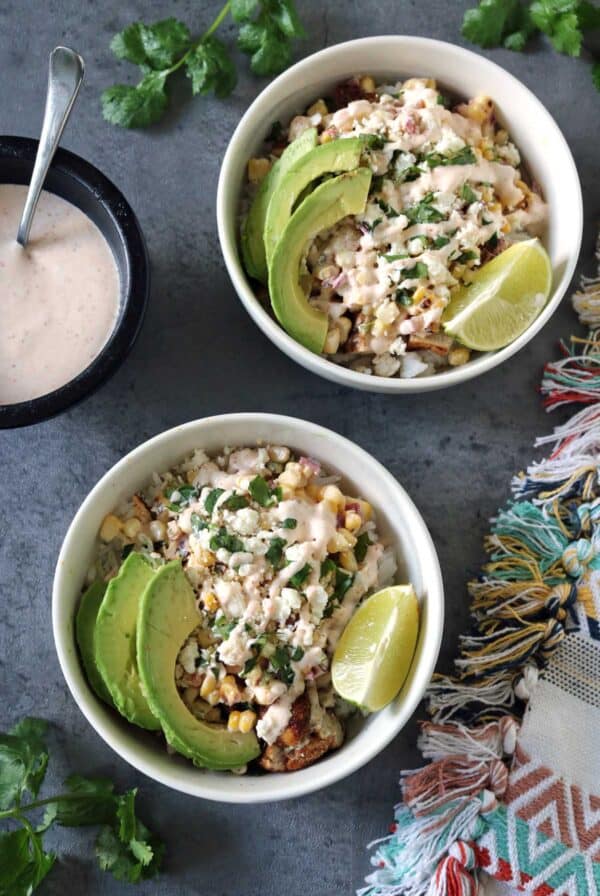 Two bowls of Grilled Street Corn Chicken Bowls topped with avocado slices, lime wedges, cheese, and creamy dressing sit on a gray surface, accompanied by a colorful cloth, fresh cilantro, and extra dressing on the side.