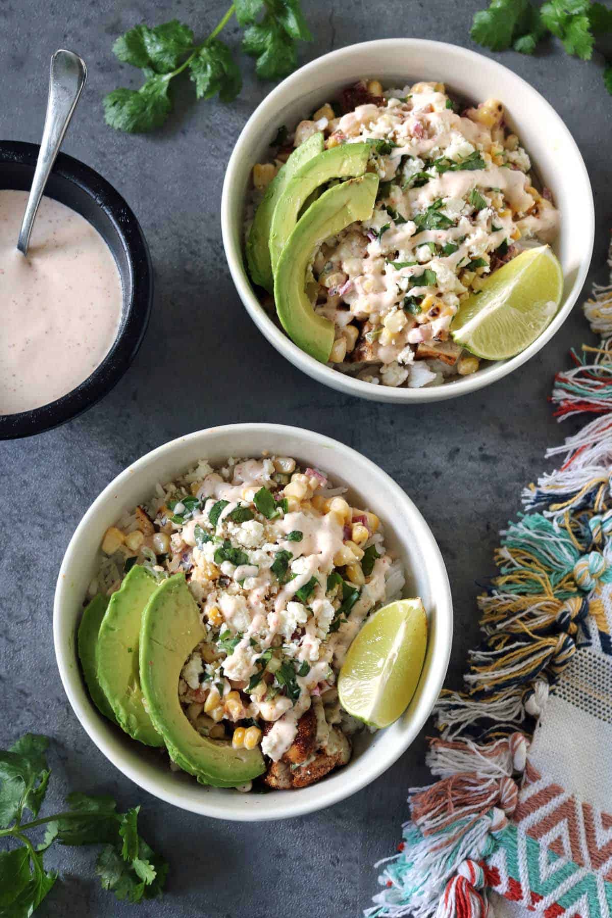 Two bowls of Grilled Street Corn Chicken Bowls topped with avocado slices, lime wedges, cheese, and creamy dressing sit on a gray surface, accompanied by a colorful cloth, fresh cilantro, and extra dressing on the side.