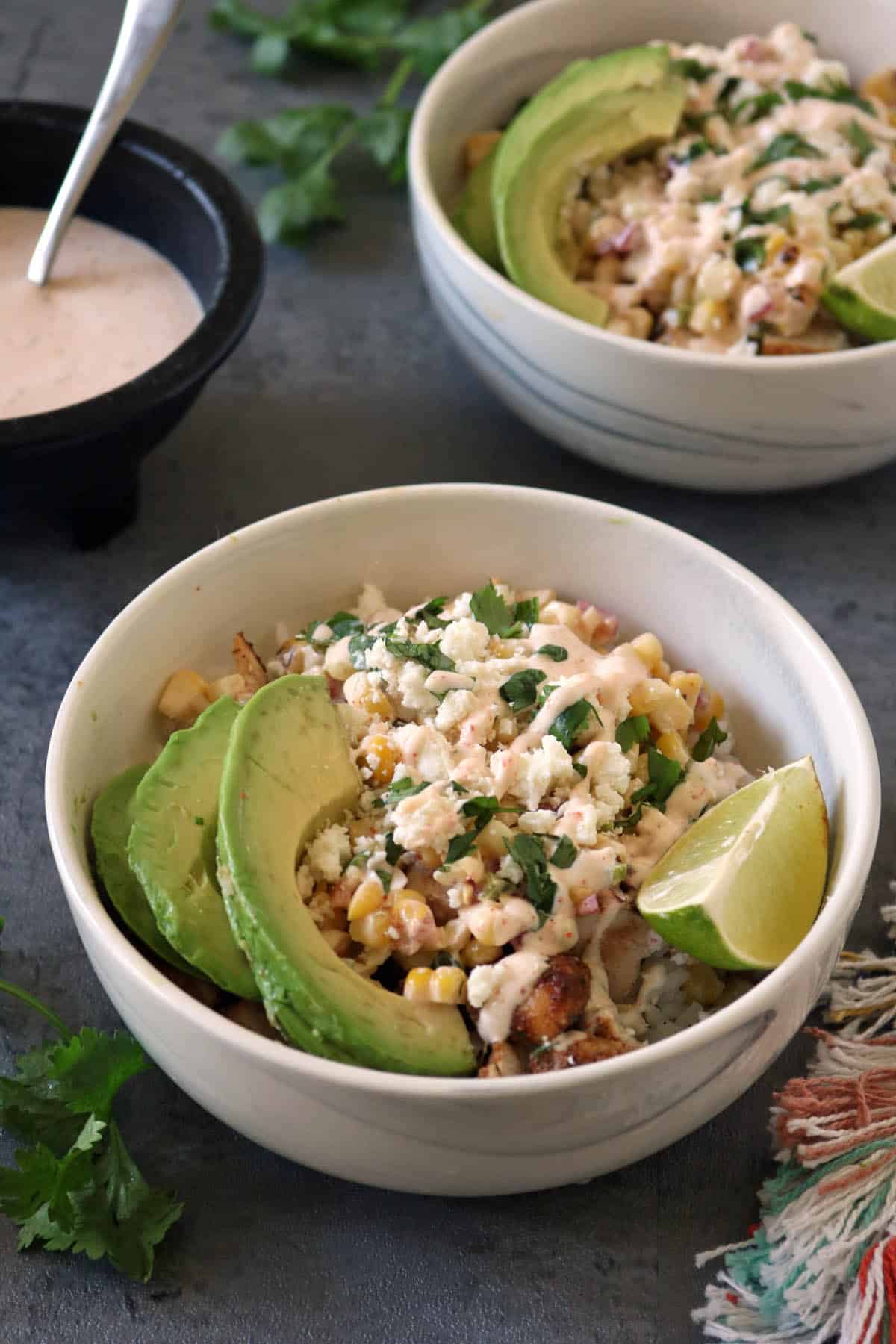 A bowl of grilled corn salad topped with avocado slices, crumbled cheese, chopped herbs, and a lime wedge, with a drizzle of creamy dressing—just like the flavors in classic Grilled Street Corn Chicken Bowls. Another bowl and dressing bowl are in the background.