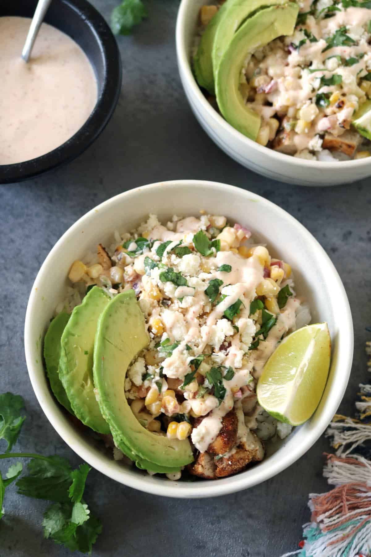 Bowl with avocado slices, charred corn, crumbled cheese, cilantro, rice, and creamy sauce, garnished with a lime wedge. Perfect for fans of Grilled Street Corn Chicken Bowls, another bowl and small sauce dish are nearby on a gray surface.