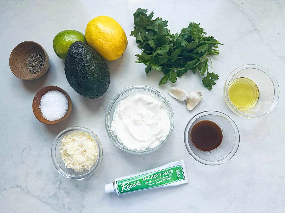 An overhead view of avocado, lemon, lime, cilantro, garlic, olive oil, black pepper, salt, grated cheese, anchovy paste, yogurt, and Worcestershire sauce on a light surface.