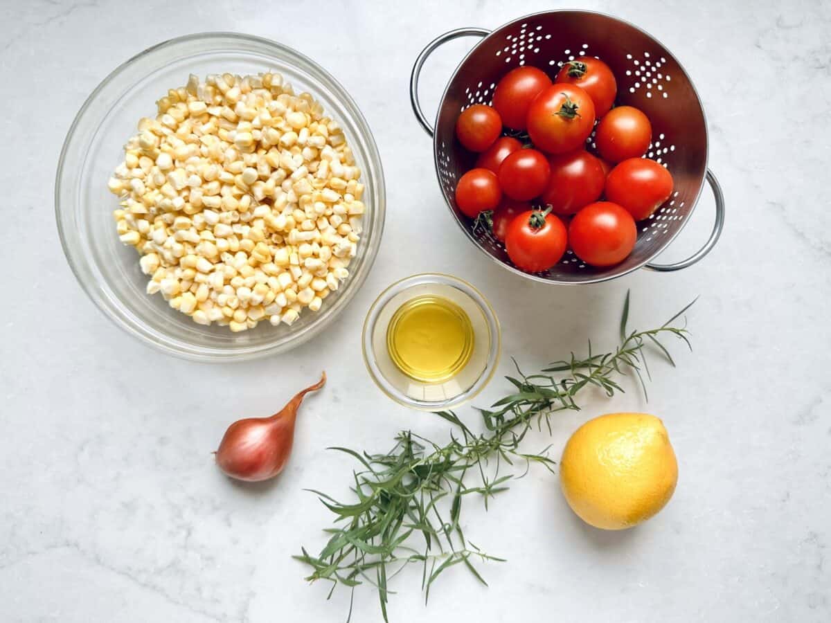A glass bowl of corn kernels, a metal colander with cherry tomatoes, a shallot, a glass cup of olive oil, fresh rosemary, and a lemon on a white countertop.