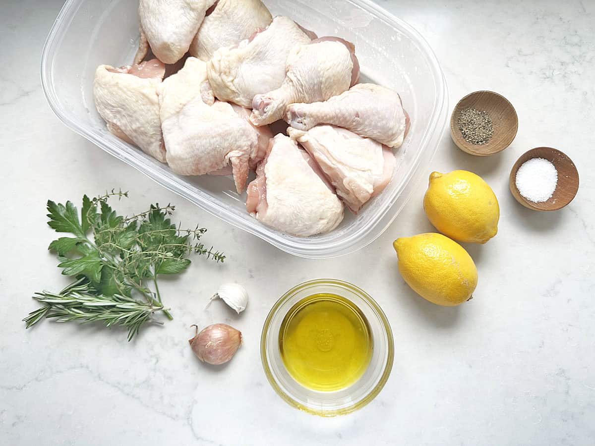 Raw chicken pieces in a plastic container, two lemons, olive oil in a bowl, a shallot, garlic clove, mixed fresh herbs, and small bowls of salt and pepper are arranged on a white surface—everything you need to create Foolproof French Grilled Chicken.