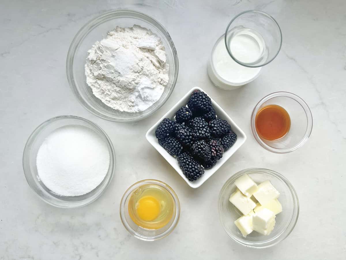 Seven ingredients on a white surface: flour, sugar, an egg, blackberries, milk, butter cubes, and vanilla extract, each in a separate bowl or glass.
