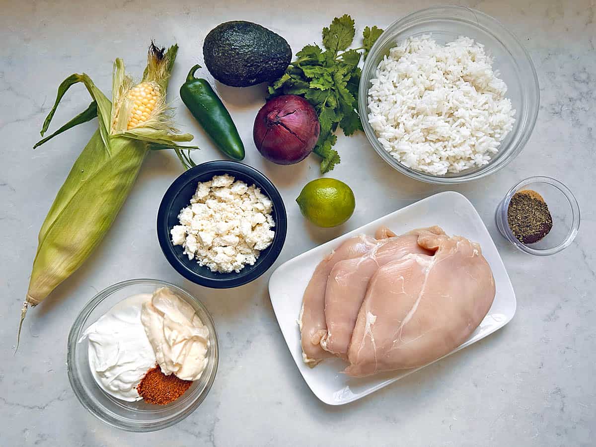 Ingredients on a countertop: raw chicken breasts, cooked rice, corn, avocado, jalapeño, red onion, cilantro, lime, feta cheese, spice blend, mayonnaise, and sour cream—perfect for assembling flavorful Grilled Street Corn Chicken Bowls.