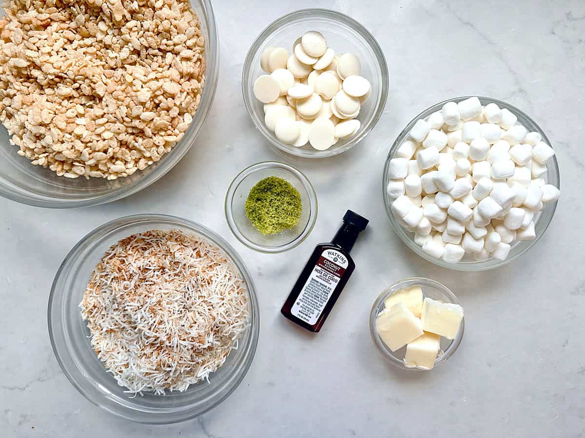 Five bowls with Rice Krispies, white chocolate, mini marshmallows, toasted coconut, ground matcha powder, plus vanilla extract and butter on a white surface.