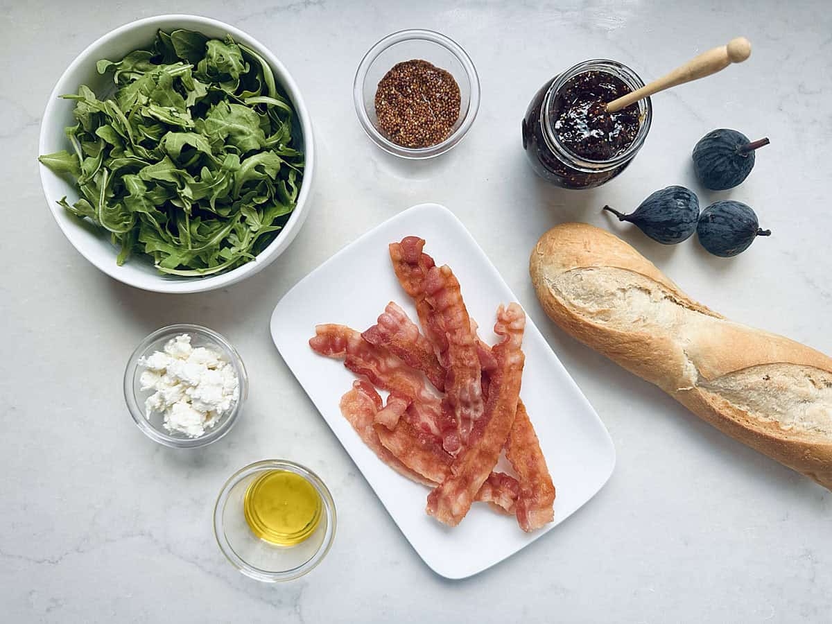 A bowl of arugula, a baguette, cooked bacon, crumbled cheese, olive oil, grainy mustard, fig jam, and fresh figs are artfully arranged on a white countertop—everything you need to create delicious Bacon Fig and Arugula Sandwiches.