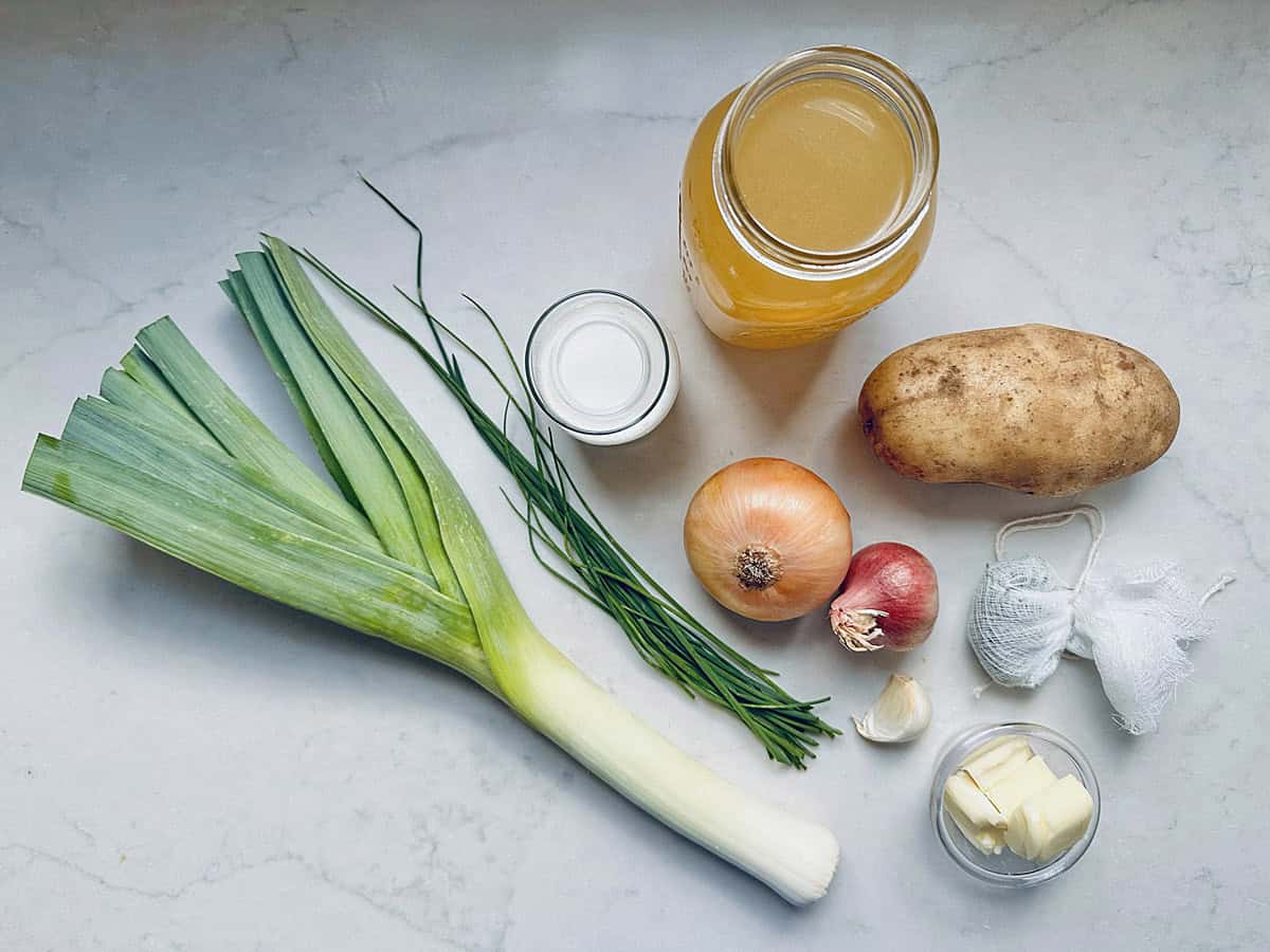A leek, chives, small jar of cream, mason jar of broth, russet potato, yellow onion, shallot, garlic cloves, butter, and a bundled herb sachet are artfully arranged on a white surface—capturing the essential ingredients for a comforting homemade Potato Leek Soup.