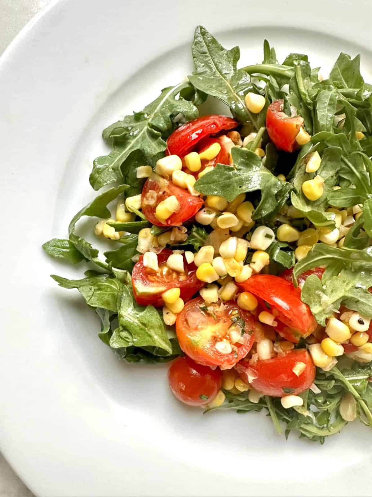 A Corn and Tomato Salad with Lemon-Tarragon Vinaigrette featuring arugula, cherry tomatoes, and corn kernels is served on a white plate.