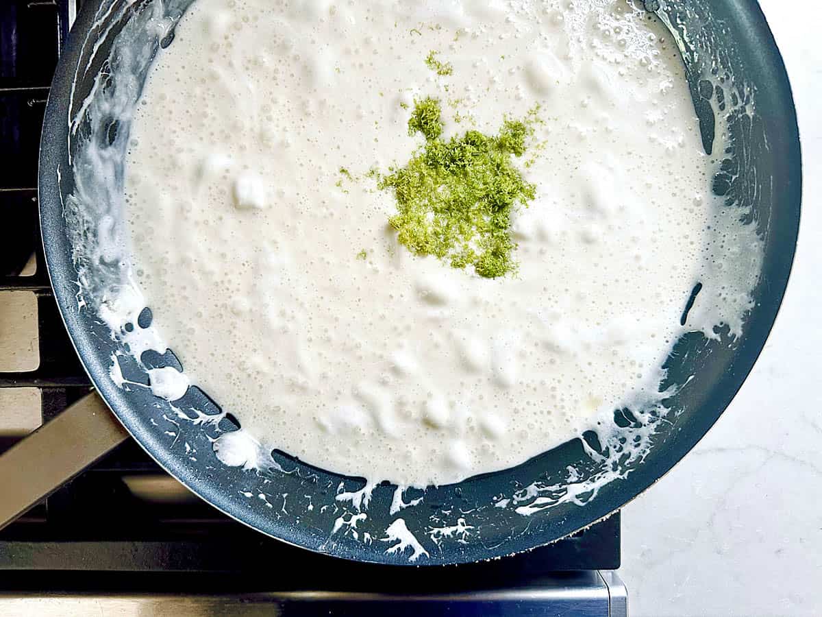 A pan on a stove contains a bubbling white mixture with a small pile of green zest, likely lime, in the center. Some of the mixture is splattered on the pans edges.