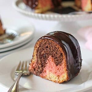 A slice of marble bundt cake with chocolate glaze sits on a white plate with a fork; the cake has swirls of chocolate, pink, and yellowโa signature touch from the galley gourmetโand more slices are on a cake stand in the background.