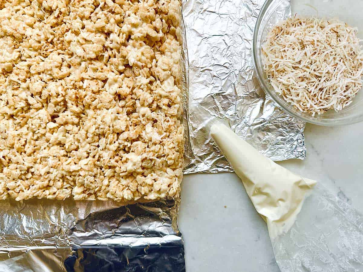 Rice Krispies treats on foil next to a piping bag filled with white icing and a glass bowl of shredded coconut on a marble surface.