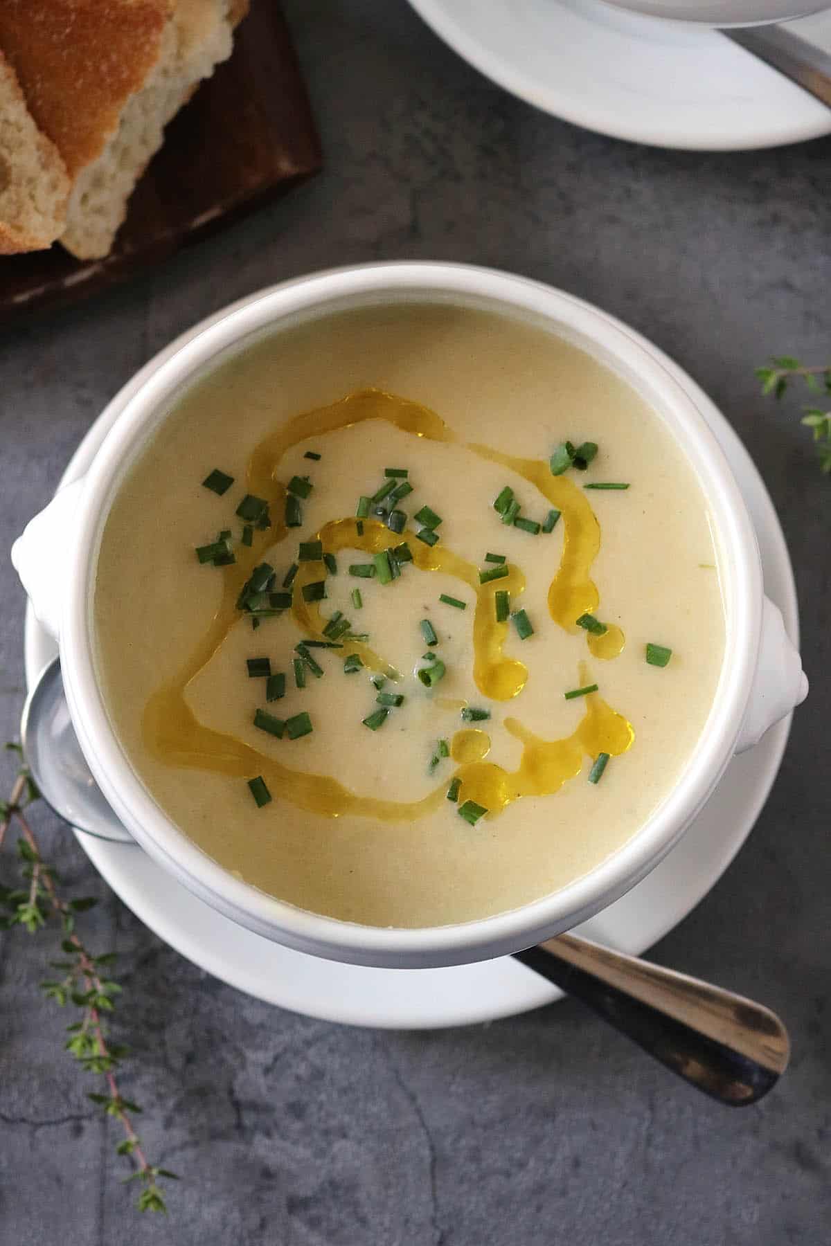 A bowl of creamy Potato Leek Soup garnished with chopped chives and a drizzle of olive oil, served with a spoon on a white saucer. Bread is visible in the background on a dark surface.