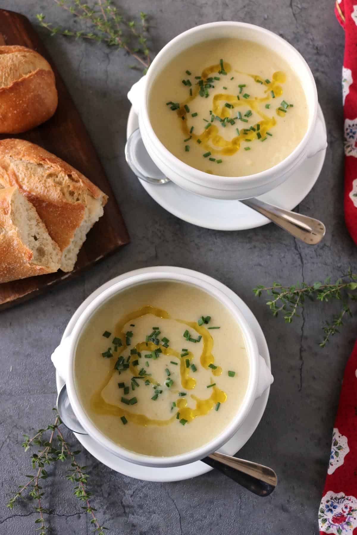 Two bowls of creamy Potato Leek Soup topped with chopped herbs and a drizzle of oil, served with metal spoons. Slices of crusty bread are on a wooden board, with sprigs of fresh herbs on a gray surface nearby.