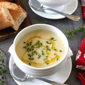 Two white bowls of creamy Potato Leek Soup topped with chopped herbs and a drizzle of oil, each on a saucer with a spoon. A loaf of crusty bread is on a wooden board nearby. Fresh herbs and a red napkin decorate the table.