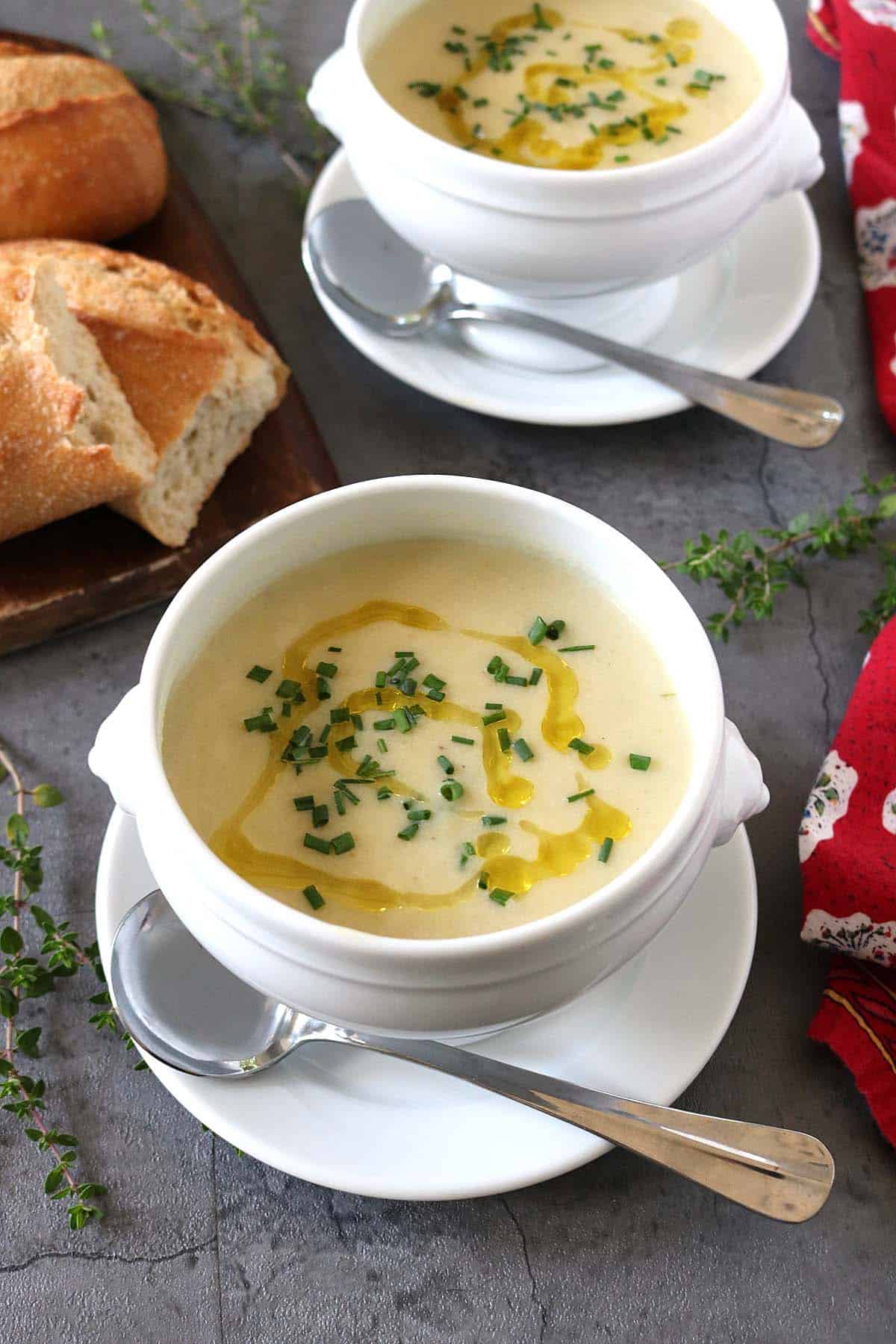 Two white bowls of creamy Potato Leek Soup topped with chopped herbs and a drizzle of oil, each on a saucer with a spoon. A loaf of crusty bread is on a wooden board nearby. Fresh herbs and a red napkin decorate the table.