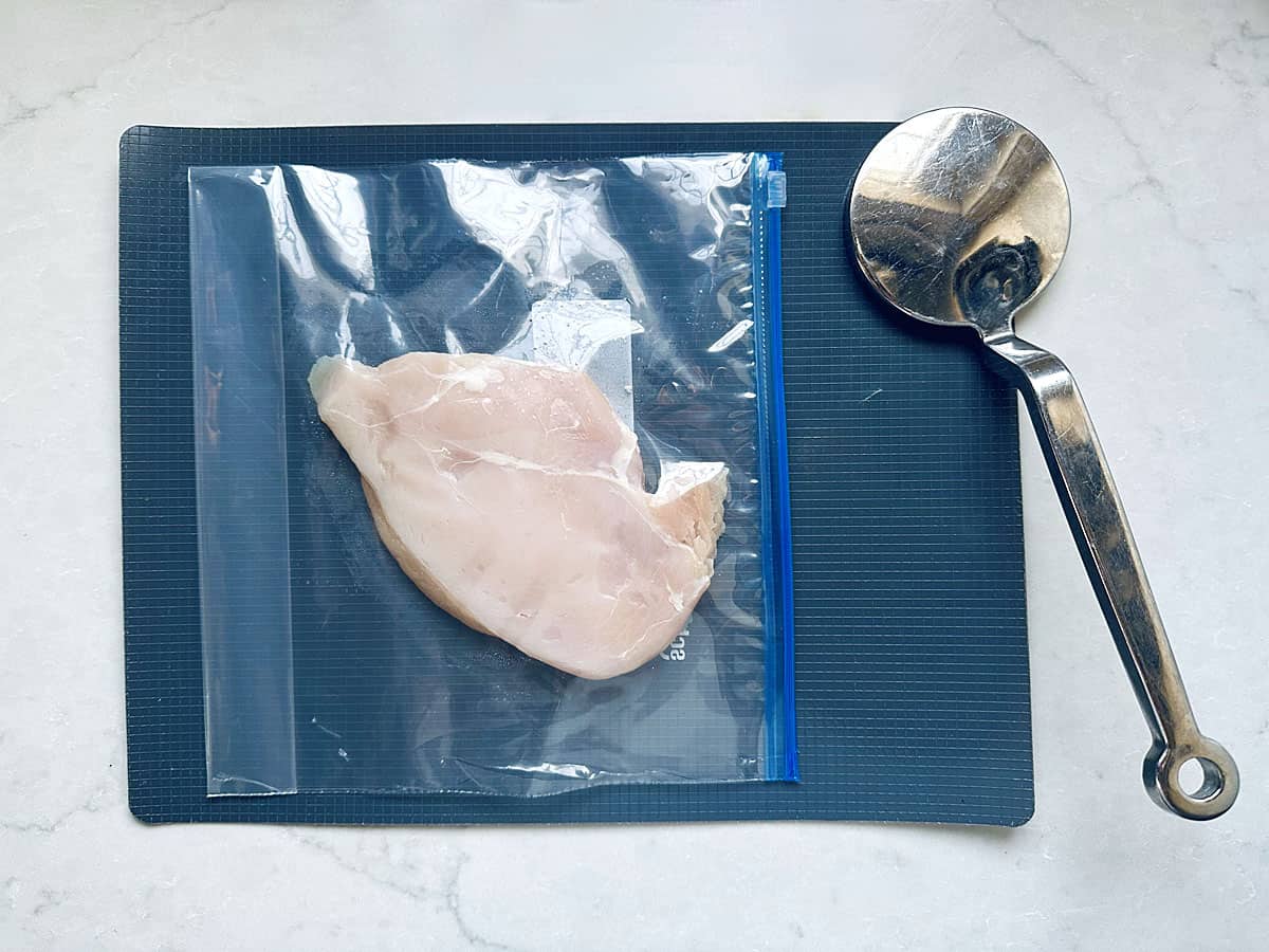 A raw chicken breast in a resealable plastic bag on a dark cutting board, next to a metal kitchen mallet on a white countertop—ready to be tenderized for Grilled Street Corn Chicken Bowls.