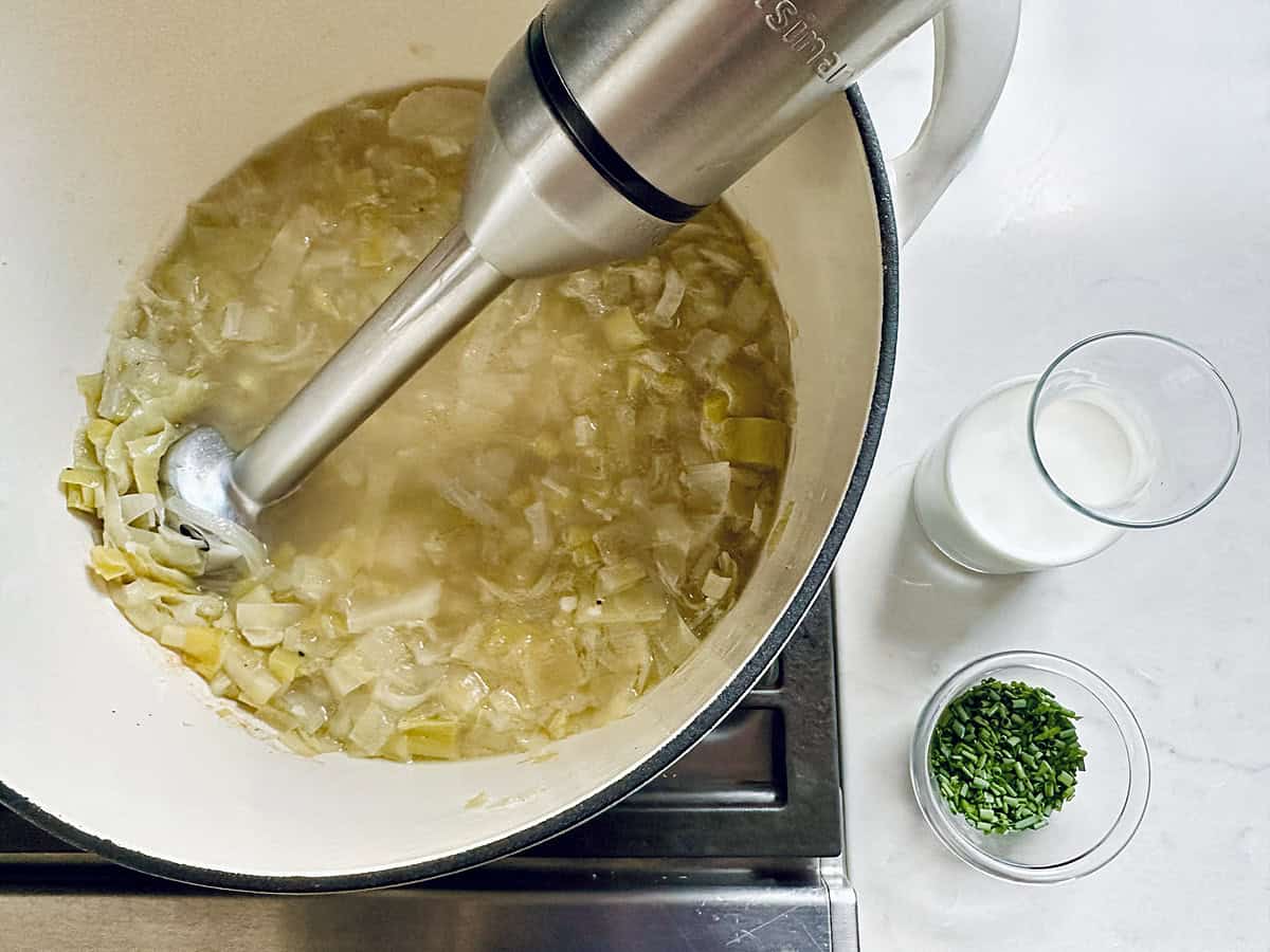 An immersion blender purees cooked leeks and broth in a pot, creating the base for classic Potato Leek Soup. Nearby on the counter are two small glasses, one with cream and one with chopped herbs, ready to be added.